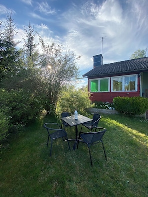 A red building with a chimney, surrounded by greenery and a table with chairs in the foreground.