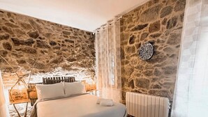 A bedroom with a stone wall, a bed with white linens, a radiator, and a small table with a lamp. - Casa as Fontes (Muíños)