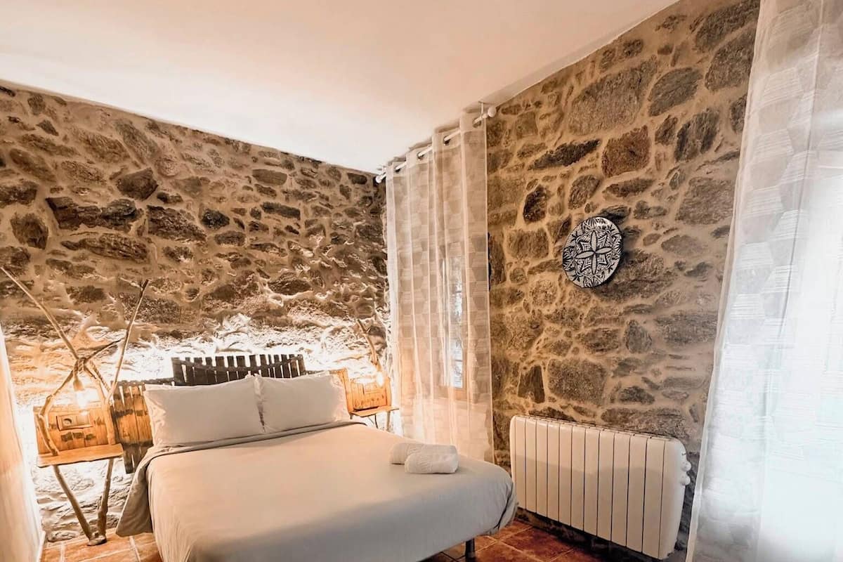 A bedroom with a stone wall, a bed with white linens, a radiator, and a small table with a lamp.