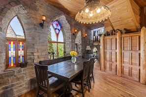 A stone-walled dining area with a wooden table and chairs, a stained glass window, and a wooden fence.