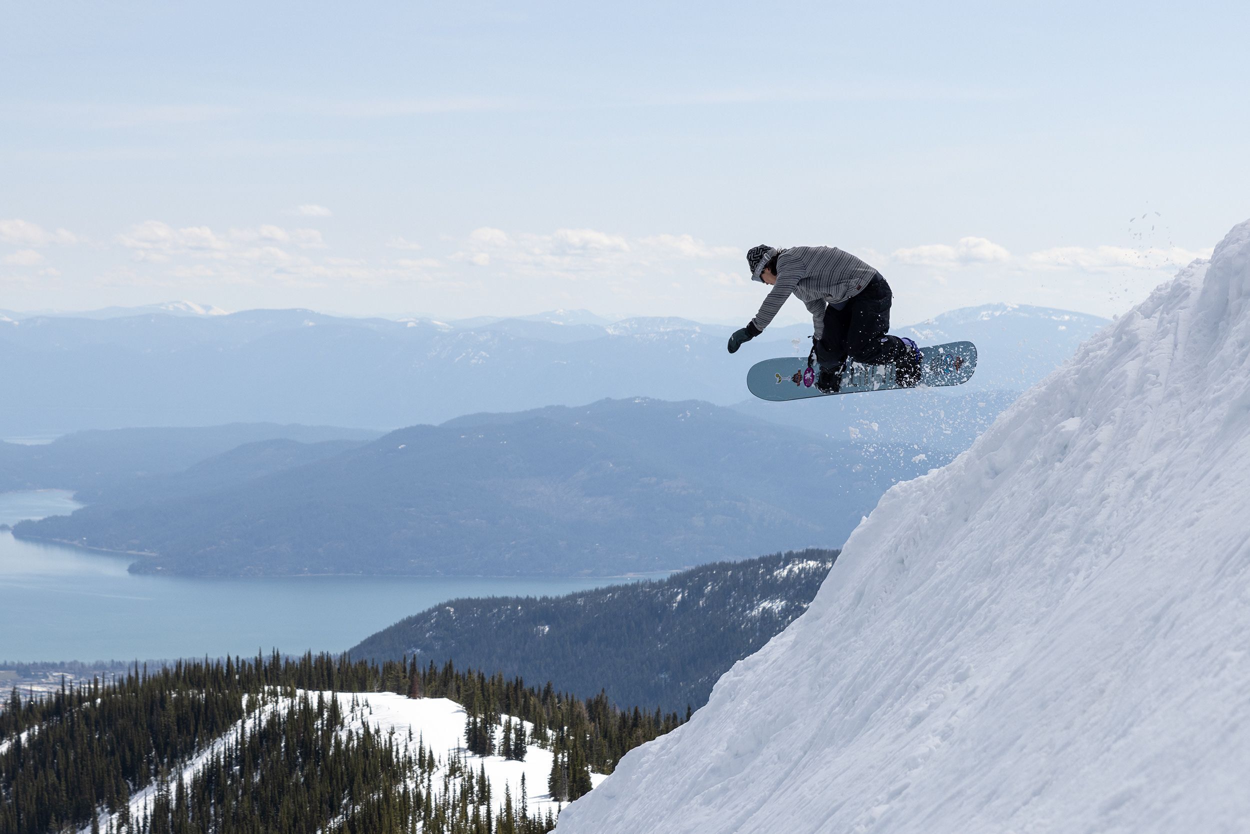 Ein Snowboarder in der Luft über einem mit Schnee bedeckten Hang, im Hintergrund ein See und Berge.