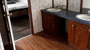 A compact bedroom with a single bed, a bathroom with two sinks and a mirror, and wooden flooring.