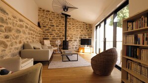 Ein Wohnzimmer mit Steinsandsteinwand, Kamin, einer Couch, einem Sessel, einem Couchtisch, einem Bücherregal und einem großen Fenster.