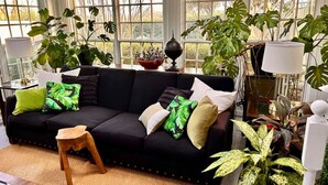 Un salon chaleureux avec un canapé noir, des coussins verts et blancs, un petit tabouret en bois et diverses plantes en pot.