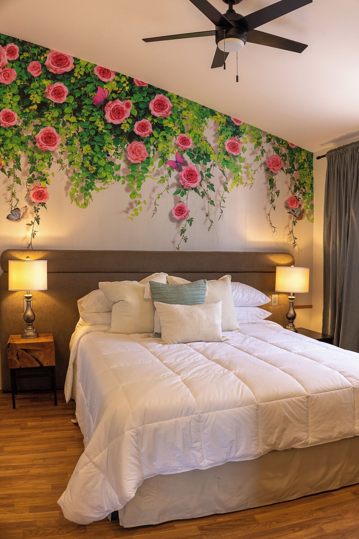 Une chambre à coucher avec un grand lit, deux lampes de chevet, un ventilateur de plafond et une fresque murale représentant des roses et de la verdure.