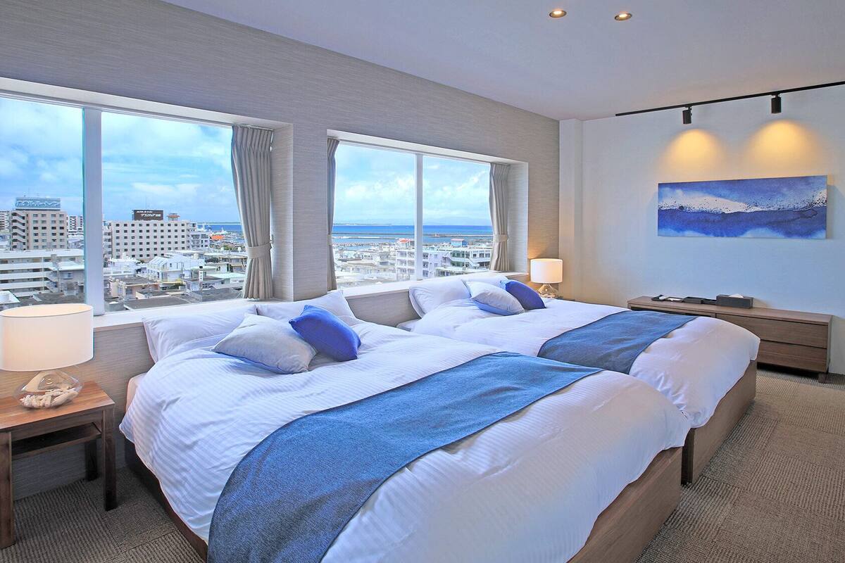 A hotel room with two beds, large windows, a city view, a painting, and a lamp.