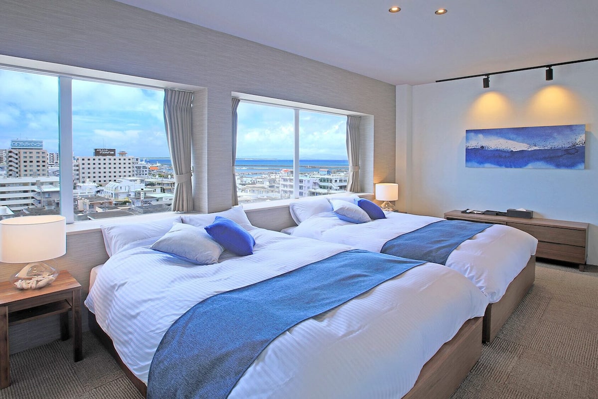 Habitación de hotel con dos camas, ventanas amplias, vista a la ciudad, un cuadro y una lámpara.
