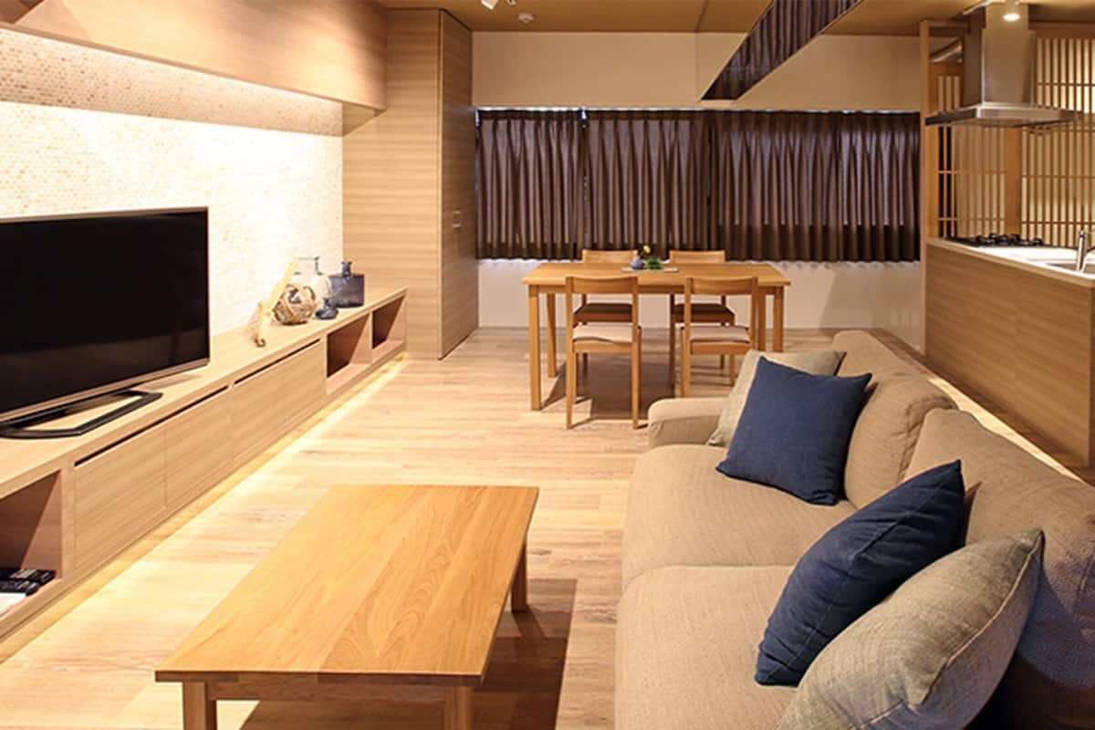 A modern living room with a flat-screen TV, wooden furniture, a dining area, and a staircase.