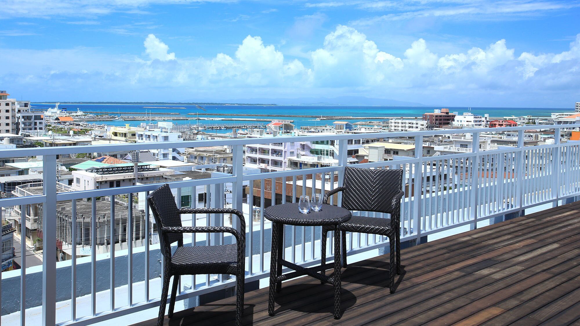 Un balcon avec des fauteuils en osier et une table, donnant sur une marina où l’on peut admirer des bateaux et des bâtiments.