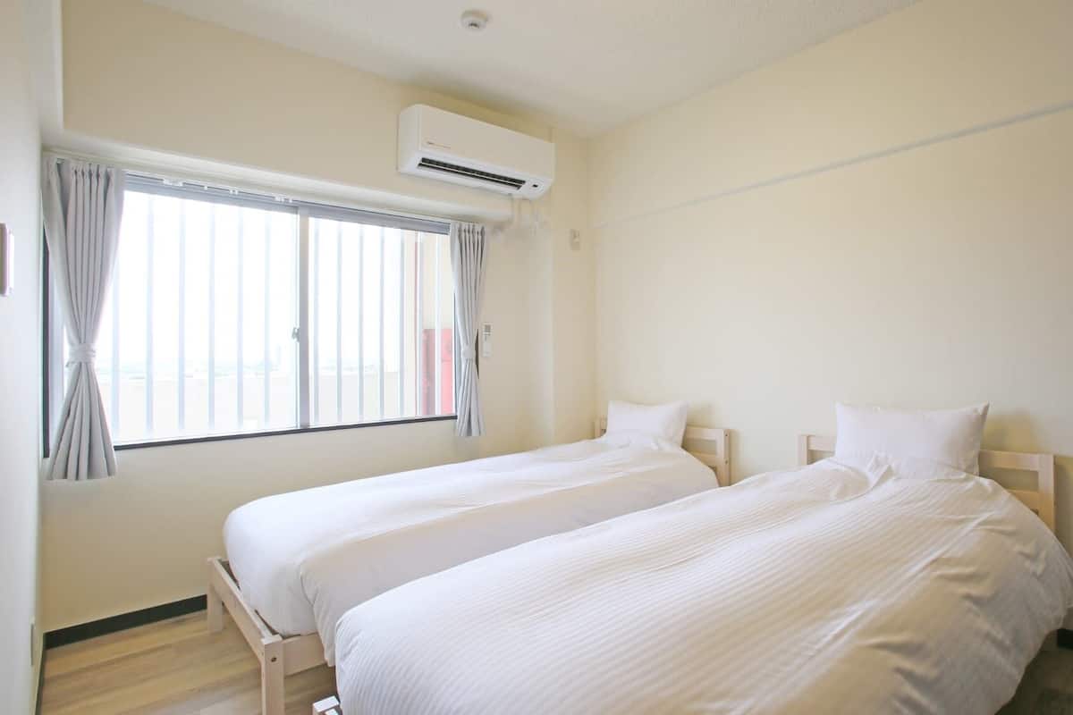 A hotel room with two single beds, a window with curtains, an air conditioning unit, and a light switch on the wall.