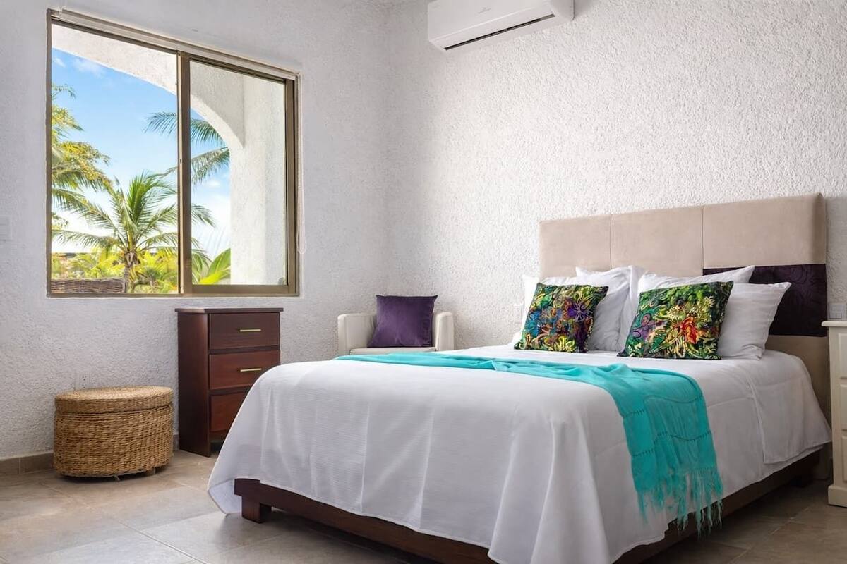 Un dormitorio con cama, ventana con vista a palmeras, un pequeño armario de madera y un taburete de mimbre.