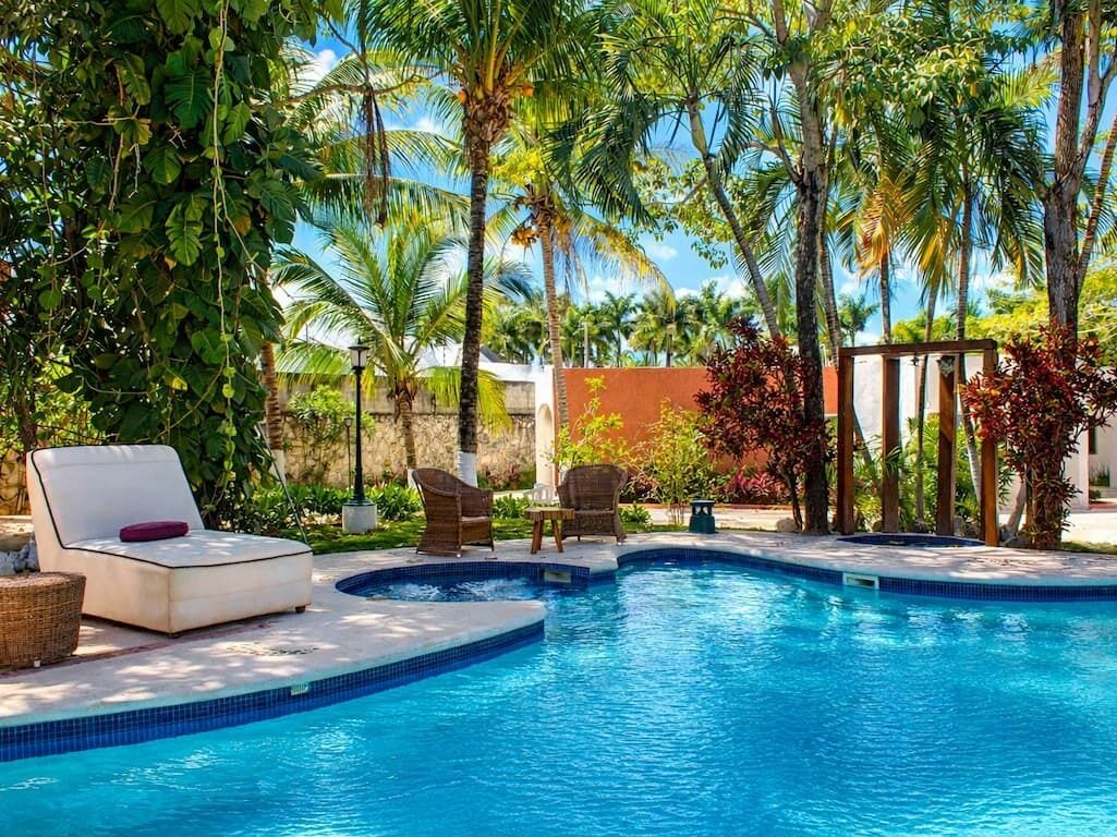 Piscina con sillas de descanso y una pequeña mesa, rodeada de palmeras y follaje.