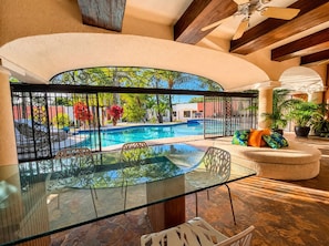 Ein von Glas umgebenes Pool-Bereich mit Blick auf ein Schwimmbecken und GrĂŒnflĂ€chen, ein runder Sitzbereich mit Kissen und ein Deckenventilator.