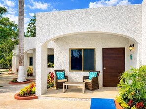 Un bâtiment à l’extérieur blanc, avec une porte sombre et deux fauteuils en osier dotés de coussins.