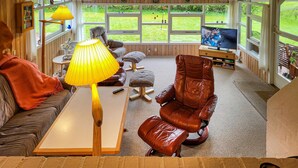 Uma sala de estar aconchegante com sofá, mesa de centro, poltrona de couro, televisão e janela com vista para um gramado.