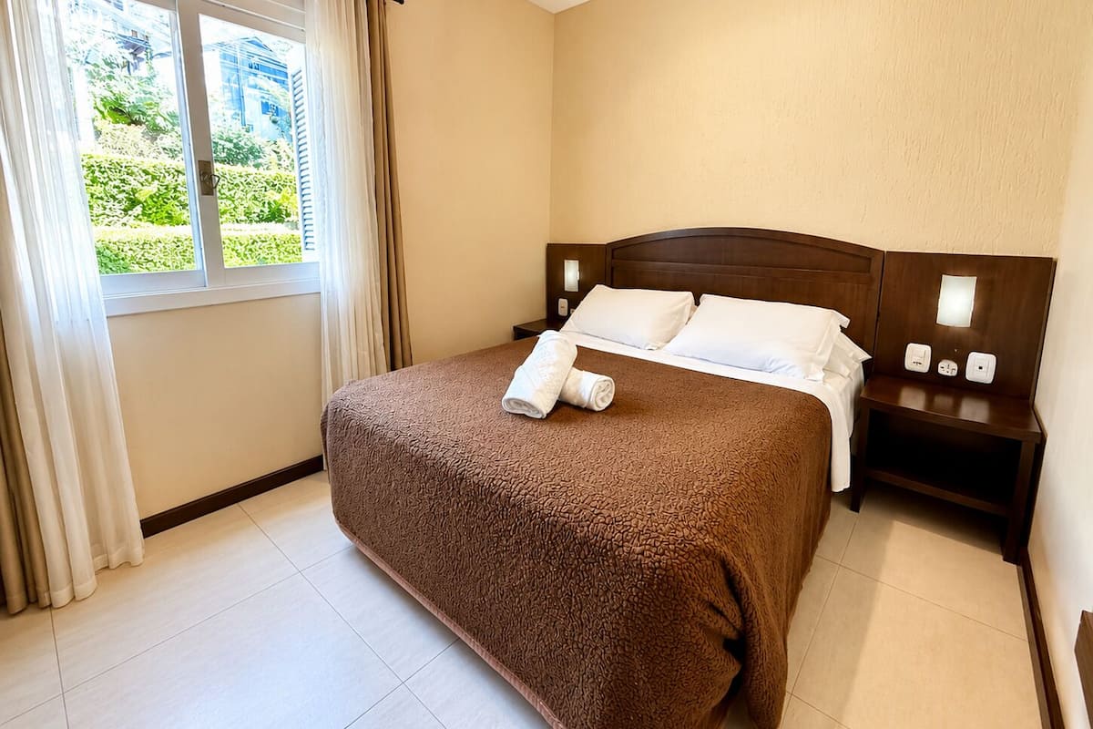 Habitación de hotel con una cama grande, una mesita de noche, una ventana con cortinas y vistas a zonas verdes.