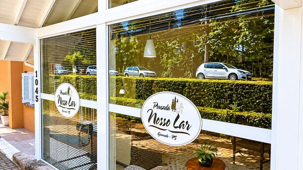 A building with large windows, a sign reading 'Pousada Nossa Lar,' and outdoor seating with a potted plant. - Pousada nosso lar (Gramado)