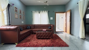 Un salon avec un canapé d’angle en cuir marron, un pouf assorti, un tapis rouge, un ventilateur de plafond et, par une porte, une vue sur un escalier.