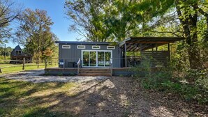 A modern cabin with a covered porch, surrounded by trees and a fenced yard.