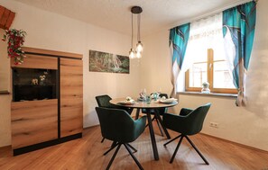 Ein Essbereich mit einem runden Tisch, vier Stühlen, einer Hängeleuchte, einem Fenster mit Vorhängen und einem an der Wand befestigten Regal mit Dekoration.