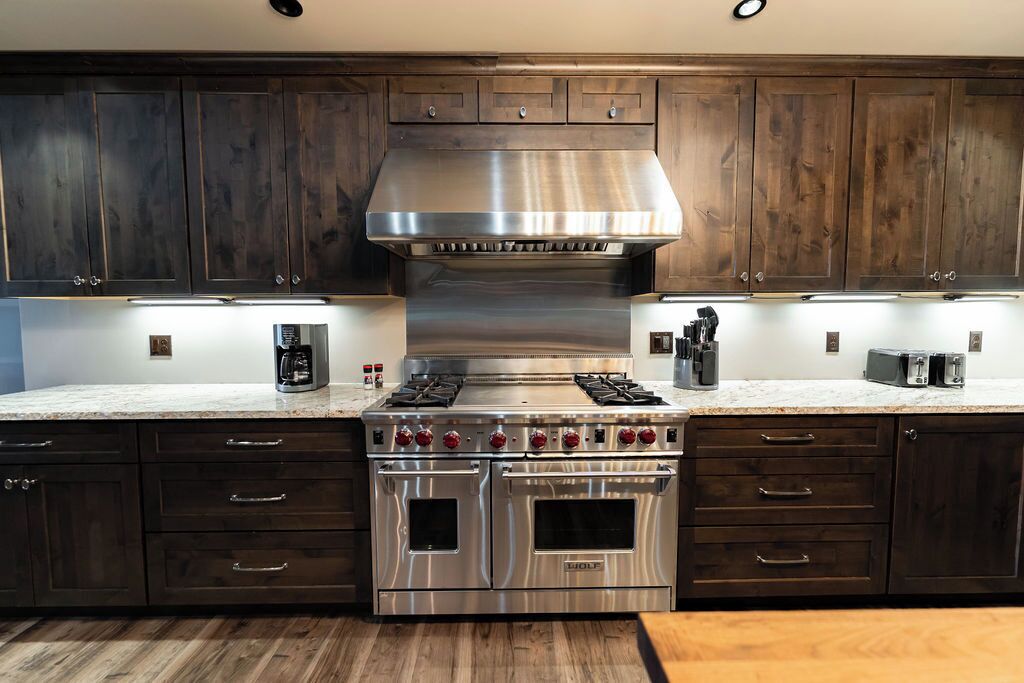 A kitchen with dark wooden cabinets, a stainless steel range hood, a double oven, and a countertop with a coffee maker and toaster.