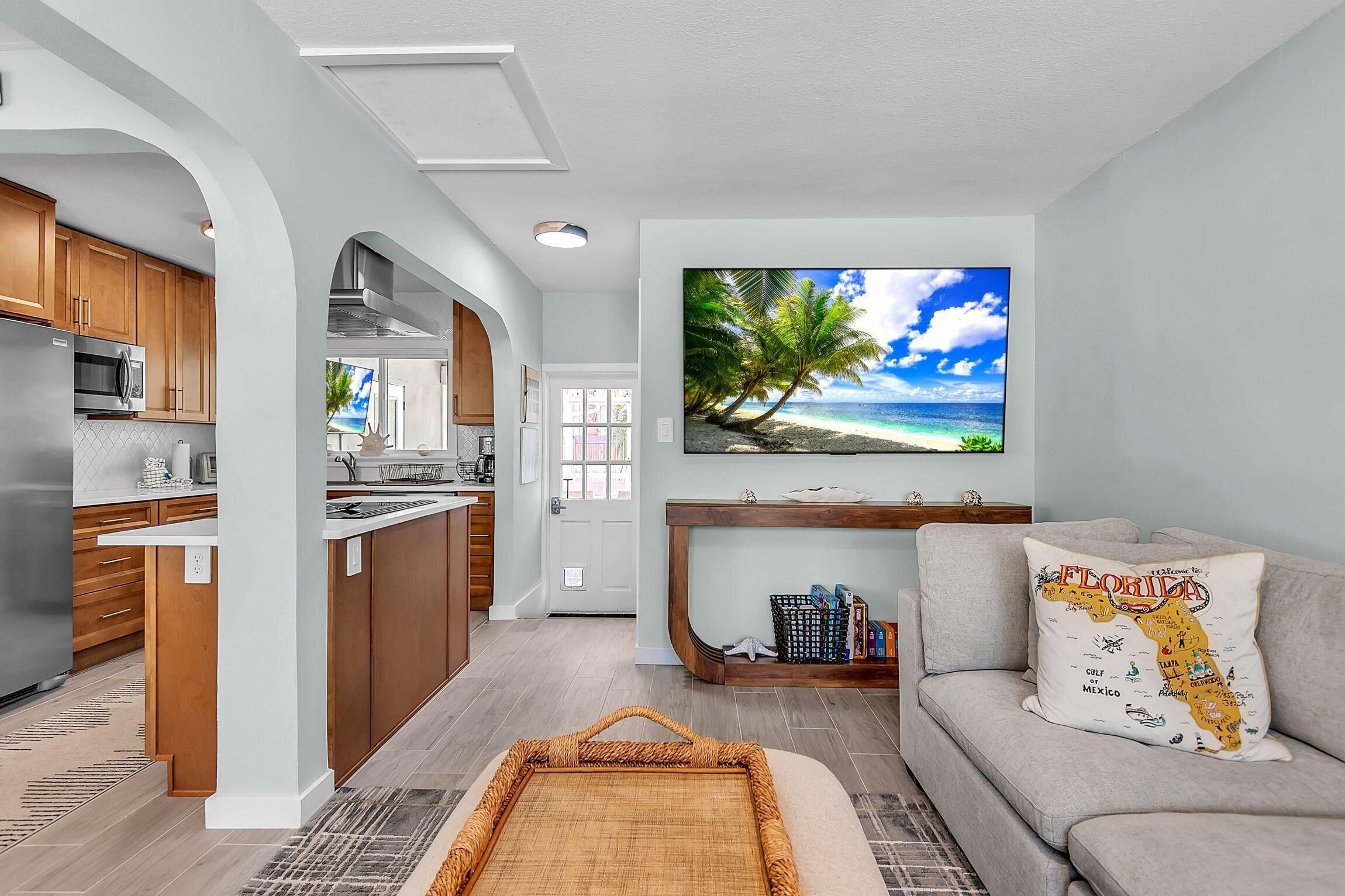 A modern kitchen with wooden cabinets and a stainless steel refrigerator, a living area with a gray sofa and a wall-mounted TV displaying a beach scene, and a small shelf with books and decorative items.
