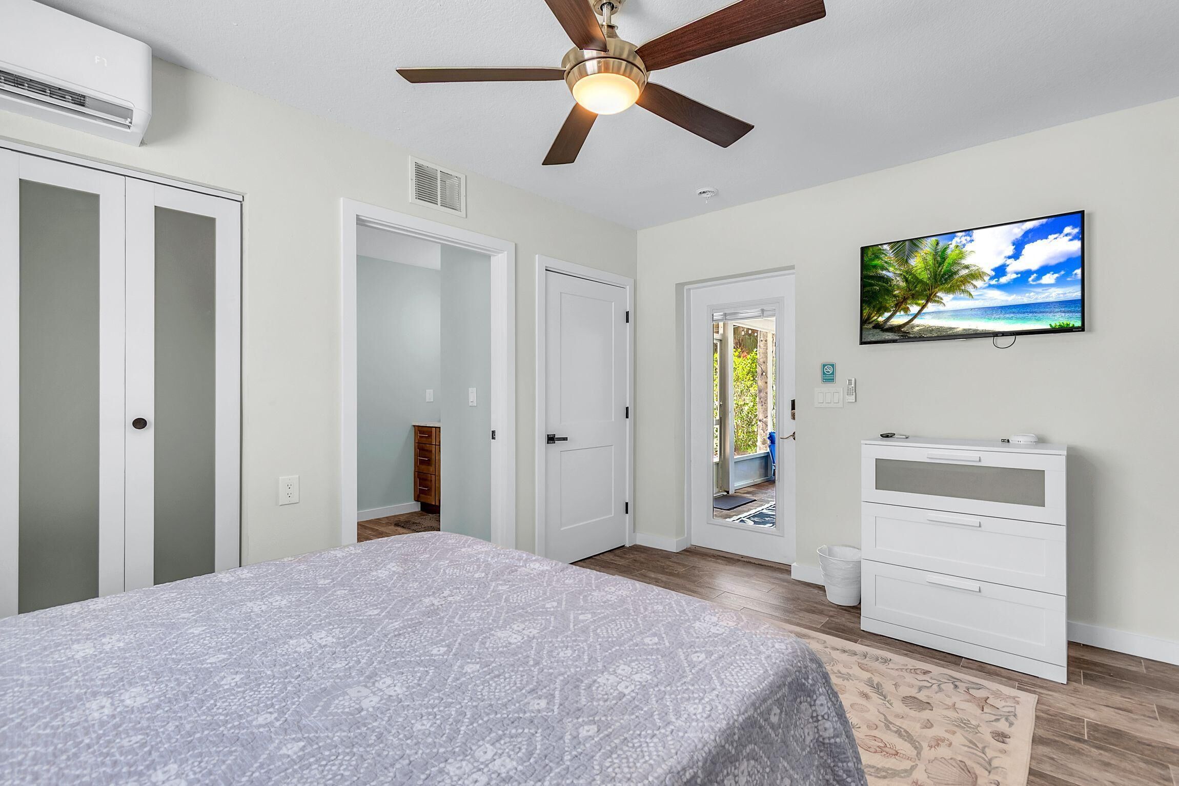 A bedroom with a bed, a ceiling fan, a TV, a dresser, and a door leading to a balcony.