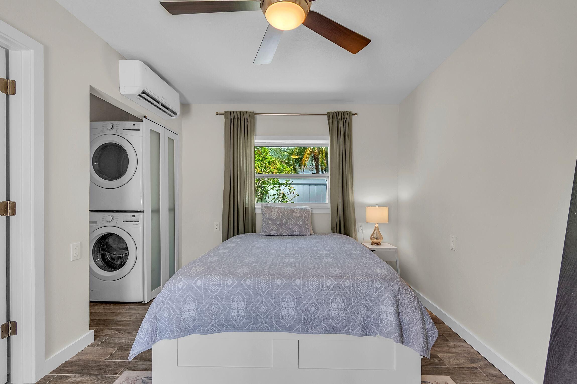 Un dormitorio con cama, ventilador de techo, aire acondicionado, ventana con cortinas y una lavadora en la habitación contigua.