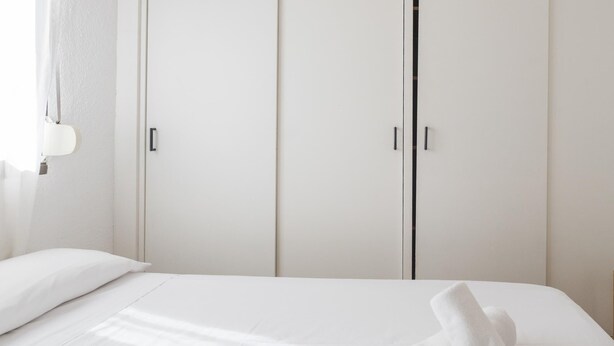 Quarto de hotel com cama, cortinas brancas e guarda-roupa de duas portas.