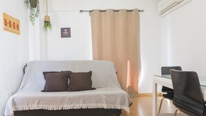 Quarto com sofá, duas almofadas, mesa de vidro, cadeira, planta pendente e cortina.