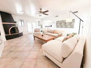 Una sala de estar amplia con chimenea, ventilador de techo, un ventanal grande y una cocina moderna al fondo.