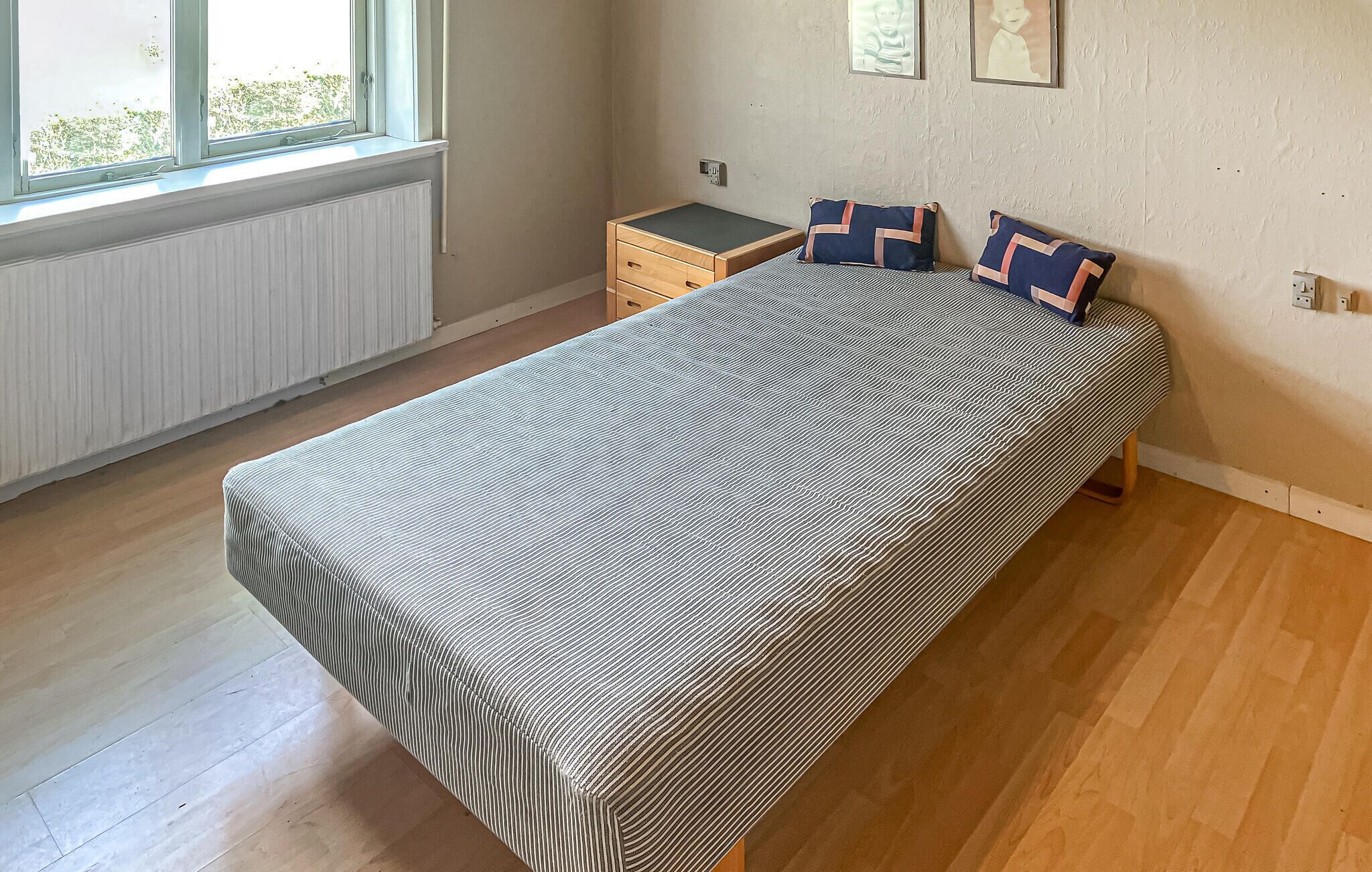 Ein Einzelbett mit gestreiftem Bettbezug, ein Nachttisch, eine Heizung und ein Fenster mit Blick ins Grüne.