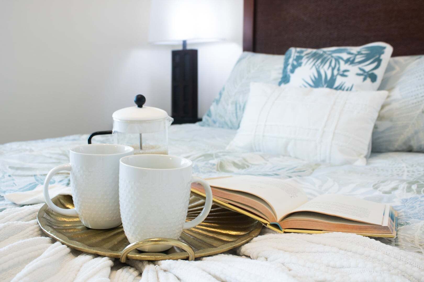 Una cama con una colcha blanca y azul estampada, una mesita de noche con una lámpara, y una bandeja con dos tazas blancas y un libro.