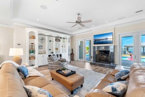 Ein großzügiges Wohnzimmer mit Kamin, Deckenventilator und Blick auf einen Pool durch große Glasfenstertüren.