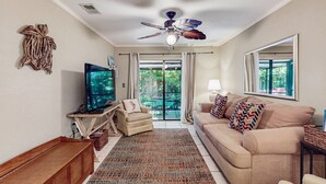 Ein Wohnzimmer mit einem beigen Sofa, einem Fernseher, einem Deckenventilator und einem großen Fenster mit Vorhängen.