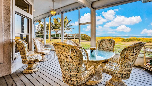 A covered outdoor seating area with wicker chairs and a glass table, overlooking a scenic landscape. - 861 S Atlantic Ave (Cocoa Beach)