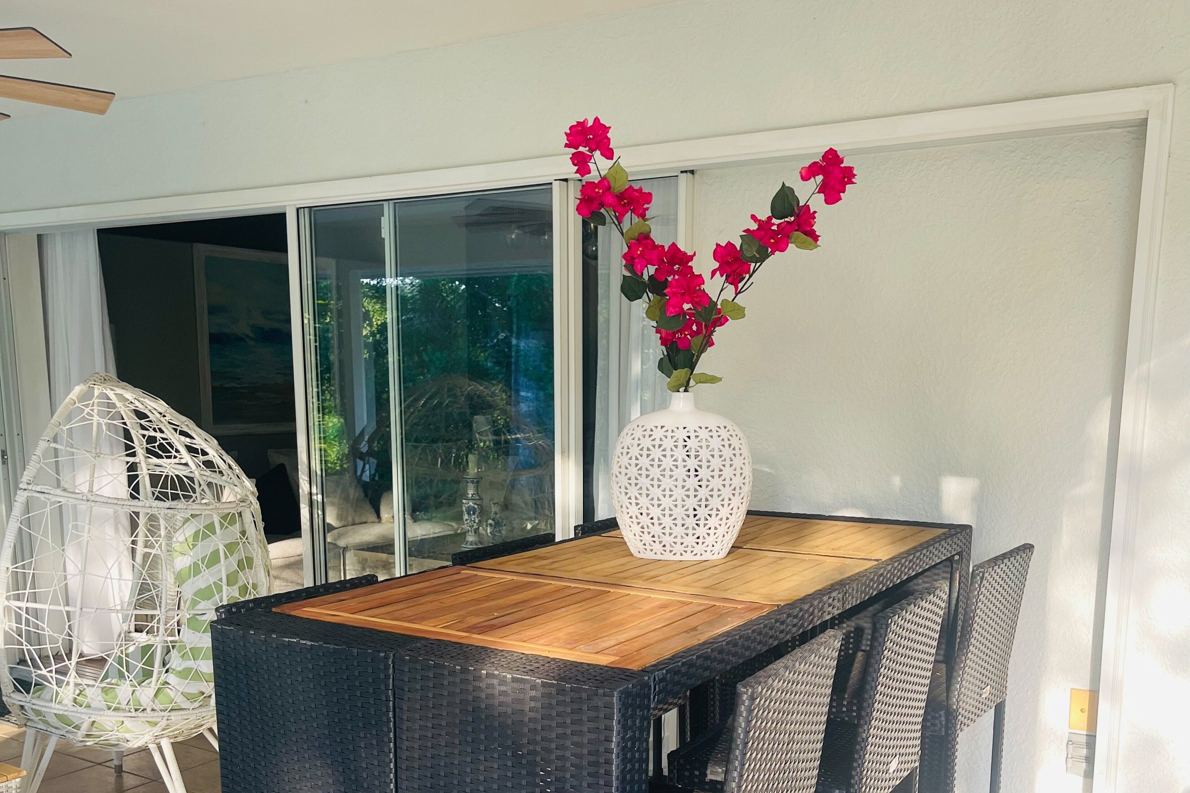 Uma área de refeições com uma mesa, cadeiras, um vaso branco com flores e um vidro que dá para uma varanda.
