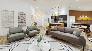 A modern living room with a sofa, armchairs, and a coffee table. There is a kitchen area with a dining table and chairs visible in the background.