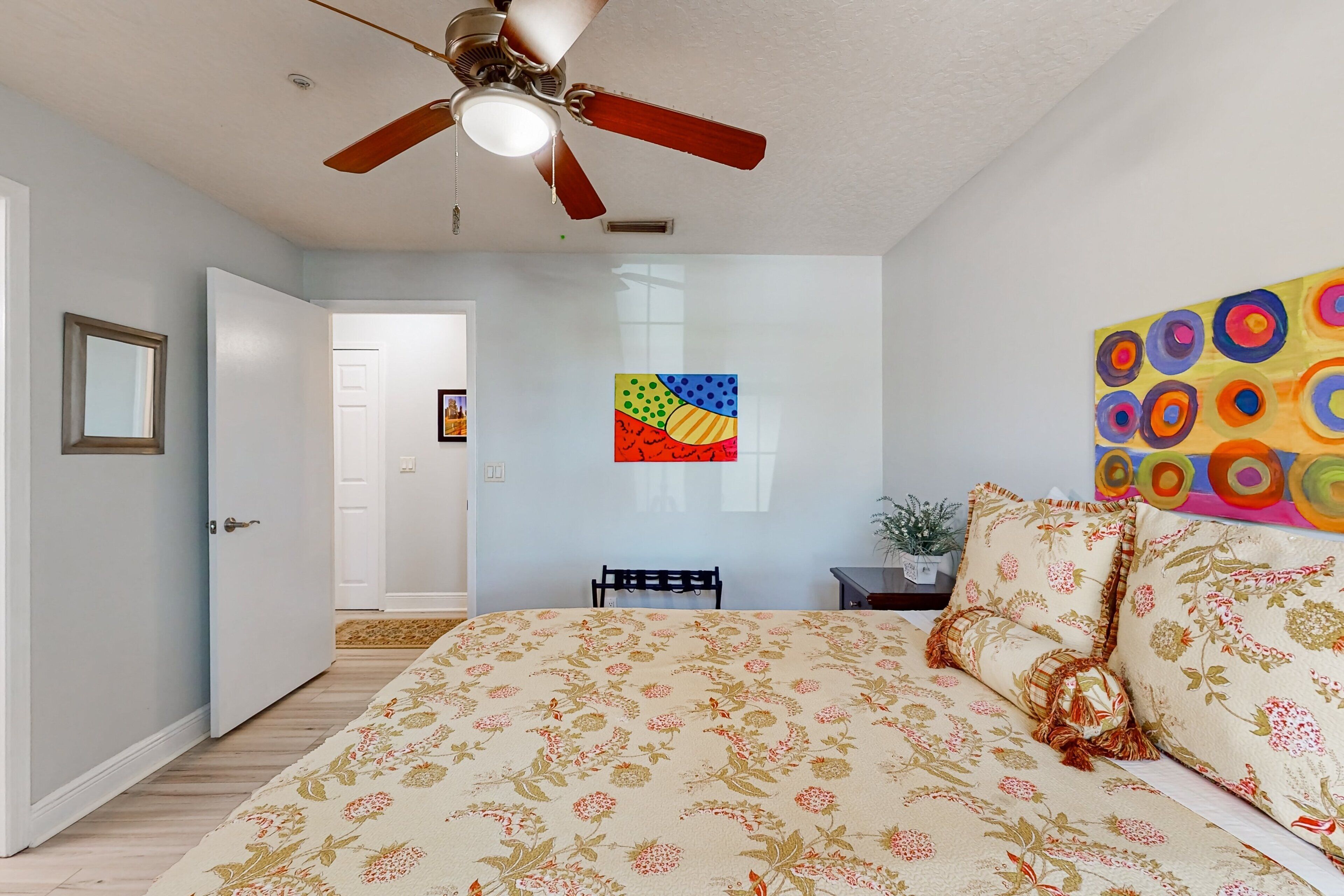 Un dormitorio con una cama, ventilador de techo, una puerta, una obra de arte en la pared y una mesita con una planta.
