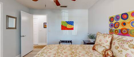 Un dormitorio con una cama, ventilador de techo, una puerta, una obra de arte en la pared y una mesita con una planta.