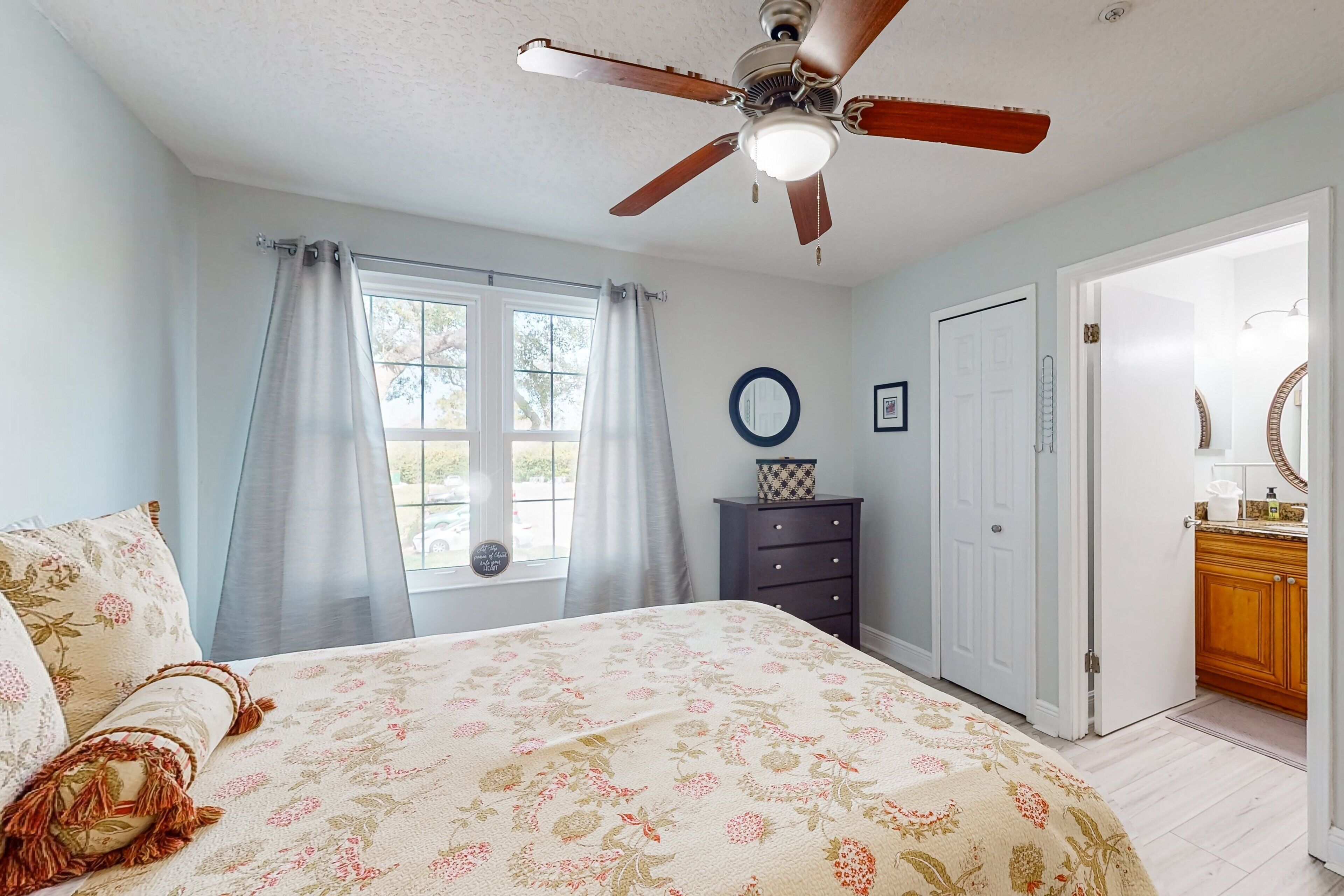 A bedroom with a bed, a ceiling fan, a window with curtains, a dresser, and a door leading to a bathroom.