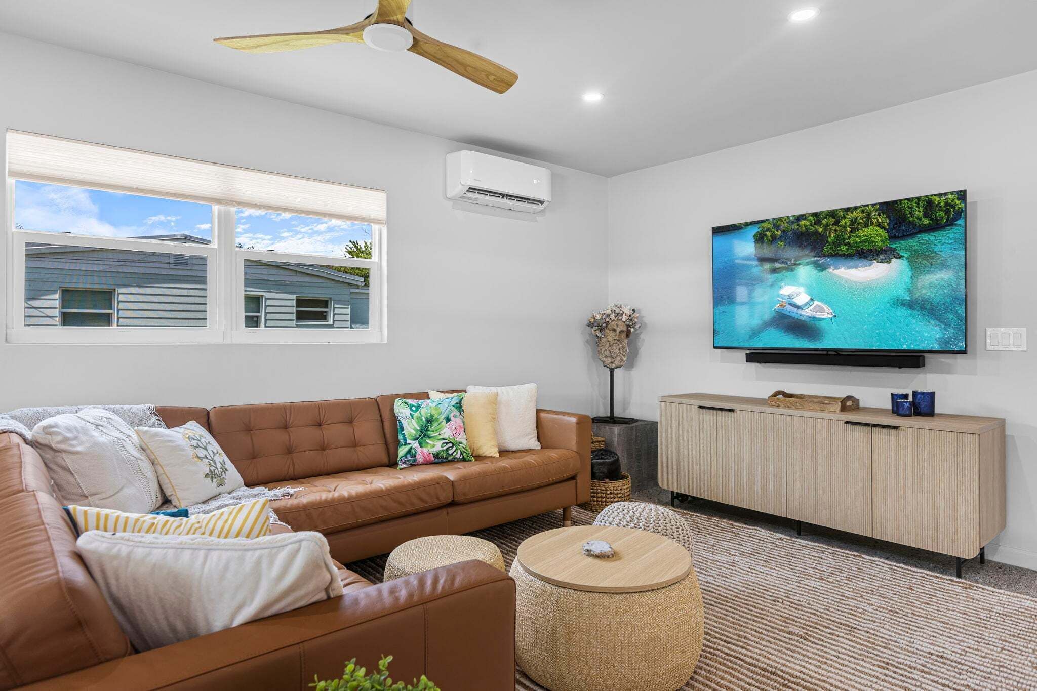 Ein Wohnzimmer mit einem braunen Ledersofa, einem runden Couchtisch, einem an der Wand montierten Fernseher und einem Deckenventilator.