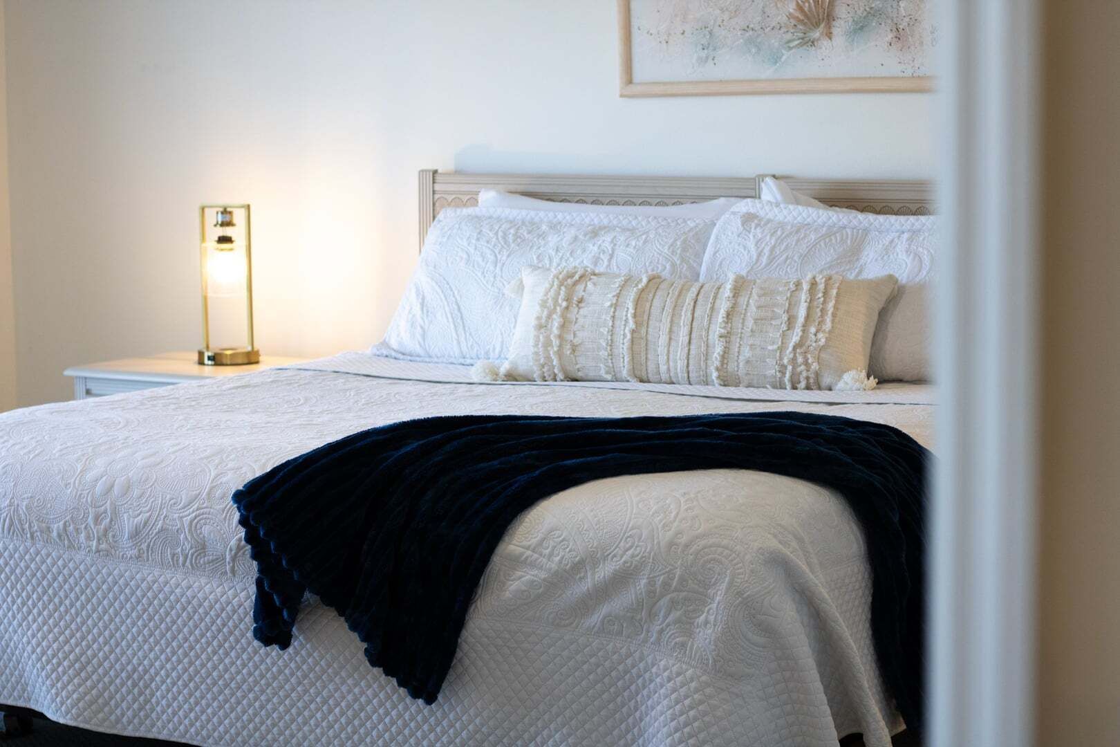 Una cama bien hecha con sábanas blancas y una manta oscura, una mesita de noche con lámpara y un cuadro en la pared.
