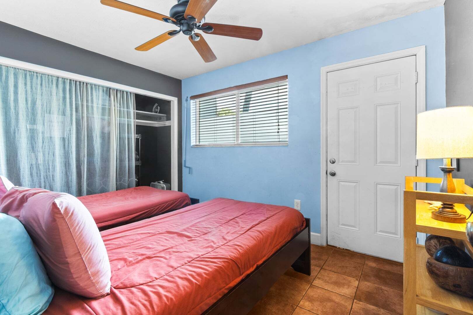 Une chambre avec deux lits, un ventilateur de plafond, une fenêtre avec des stores, une porte et une table de chevet avec une lampe.