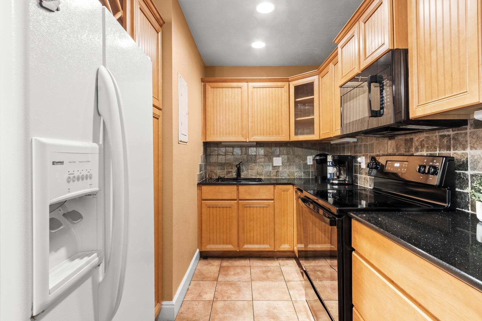 Une cuisine avec des meubles en bois, un plan de travail noir, un réfrigérateur blanc et un four encastré.