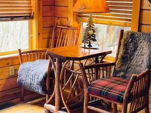 Een houten blokhut van binnen met een eettafel, stoelen, een lamp en een fauteuil met een bontdeken.