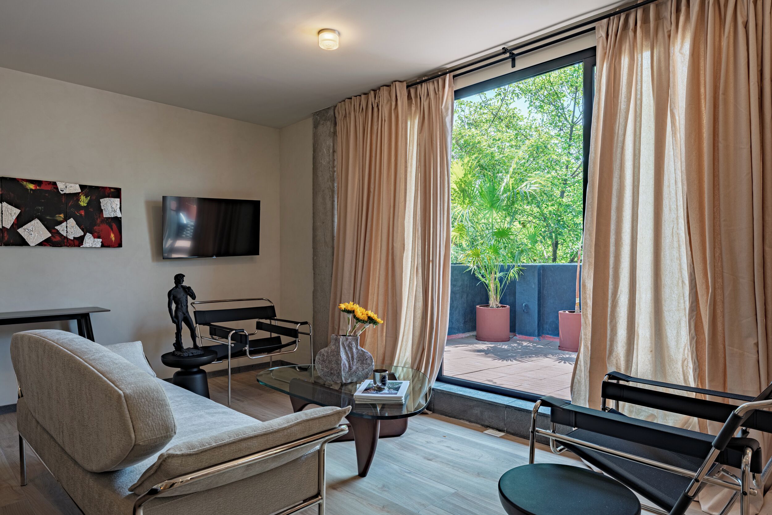 A modern living room with a sofa, a glass coffee table, a TV, and a view of greenery outside.