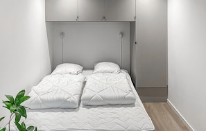 Ein Einzelbett mit weiß gesteppter Matratze und zwei Kissen, ein kleiner Nachttisch und ein wandmontiertes Schrankregal über dem Bett.