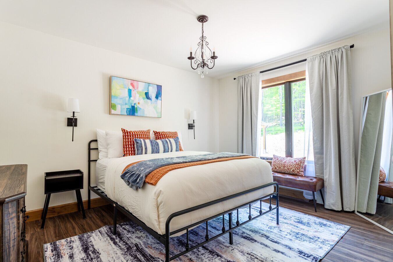 Un dormitorio con cama, mesitas de noche, candelabro, ventana con cortinas y un cuadro en la pared.