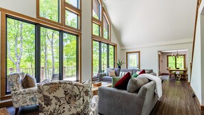 Un salon spacieux doté de grandes fenêtres, d’un canapé gris, d’un fauteuil à motifs floraux et d’un coin repas avec une table et des chaises.